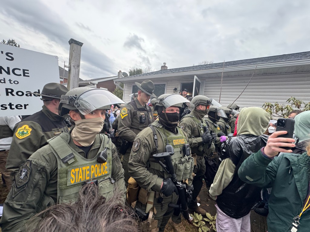 Masked VT State Police officers in riot gear stand in line between community members and the house
